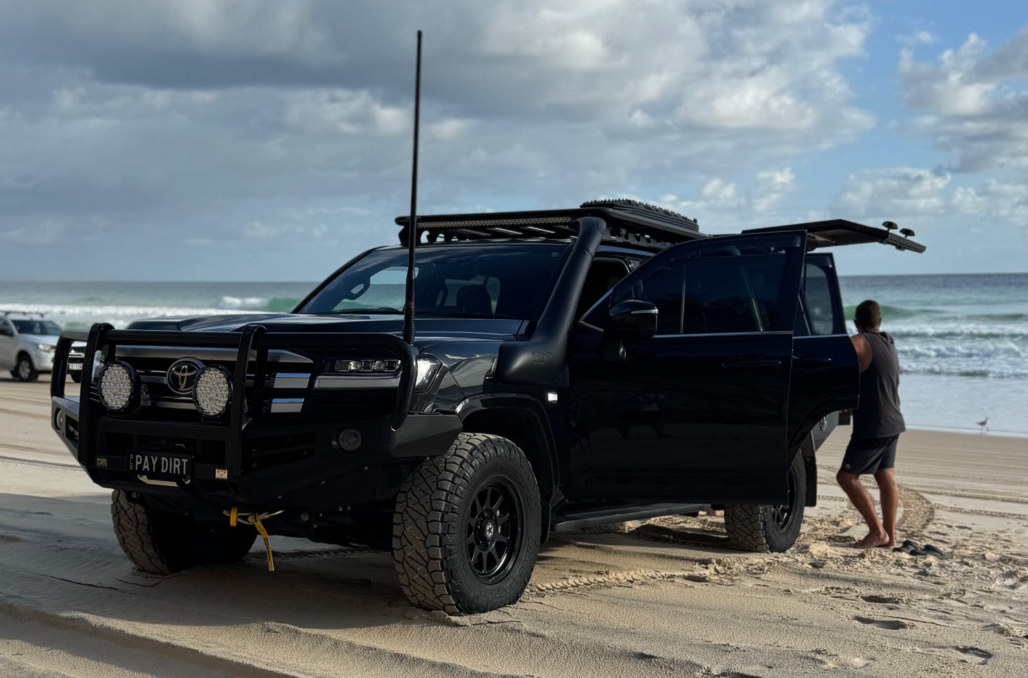 Black Toyota 300 land Cruiser on a beach with a person standing nearby